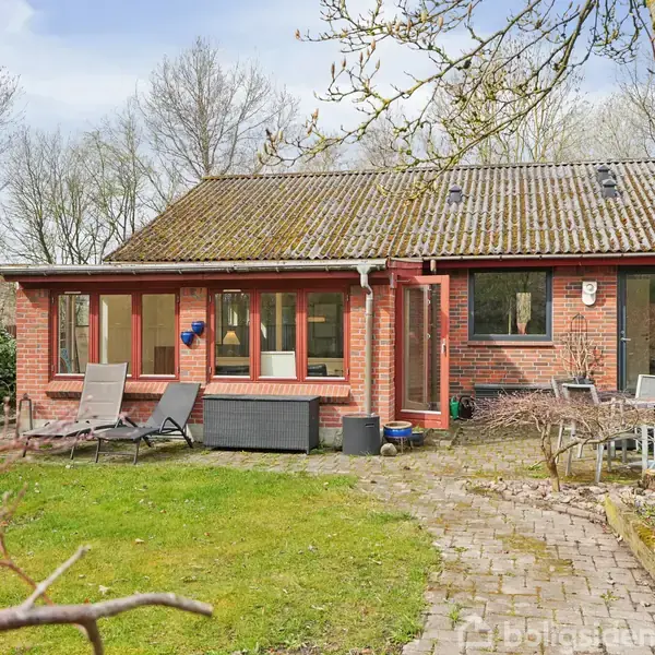Et rødt murstenshus med store vinduer. Liggestole står foran huset på en flisebelagt terrasse. Haven er grøn med træer og havemøbler. Atmosfæren er fredfyldt og naturlig.