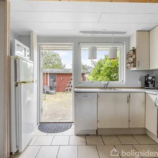 Køkken mod en åben terrassedør fører ud til en gårdhave med en rød bygning. Rummet har hvide skabe, køleskab, og en ovn. Grønne planter ses udenfor.