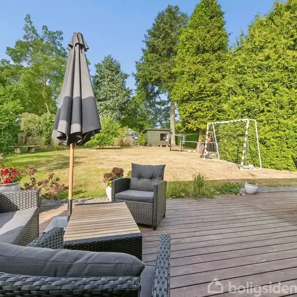 Terrassemøbler i flet står på en træterrasse med en paraply ovenover. I baggrunden ses en græsplæne med et fodboldmål og buske. Klart vejr med blå himmel.