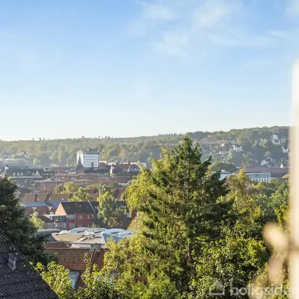 Byudsigt med grønne træer, hustage og kirketårn. I baggrunden ses en kuperet bakke under en blå himmel. Udsigten er fra et højt vindue til højre.