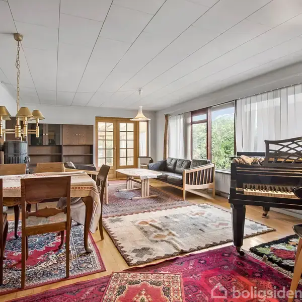Et rummeligt, velindrettet opholdsrum med et mørkt klaver i højre hjørne. Et spisebord med stole står til venstre. Store vinduer og tæpper dekorerer rummet.