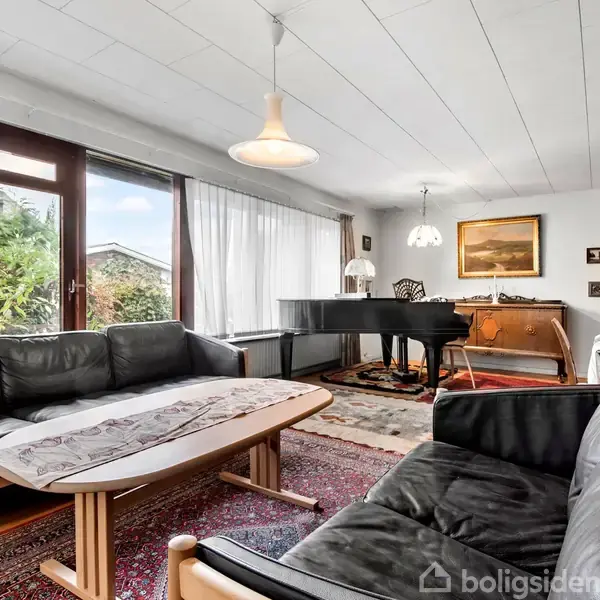 Sort læder sofagruppe står omkring et sofabord i en stue med store vinduer, der lukker dagslys ind. Et flygel står ved siden af, omgivet af møbler og kunstværker.