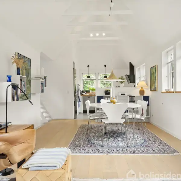 En moderne dagligstue med lyse vægge og store vinduer, møbleret med en brun lænestol, et rundt hvidt spisebord og seks stole. Et tæppe ligger centralt, omgivet af kunstværker.