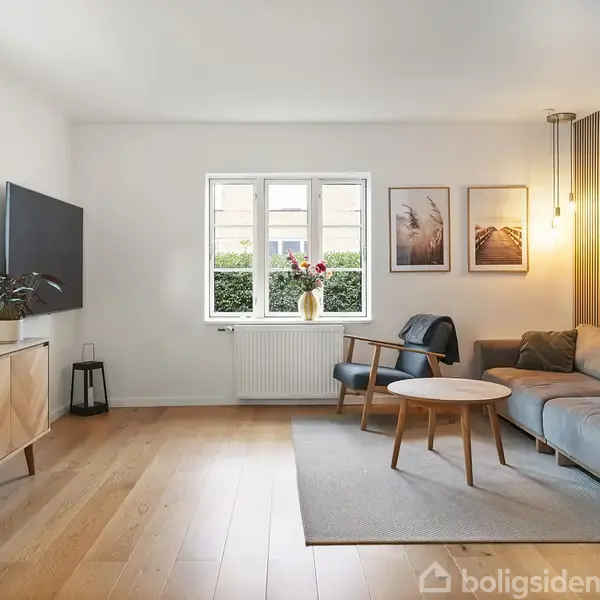 Stue med grå sofa, sofabord og armstol på tæppe. TV og planter på skænk til venstre. Tre billeder på væggen over sofaen. Vindue i midten, lysende lampe til højre.