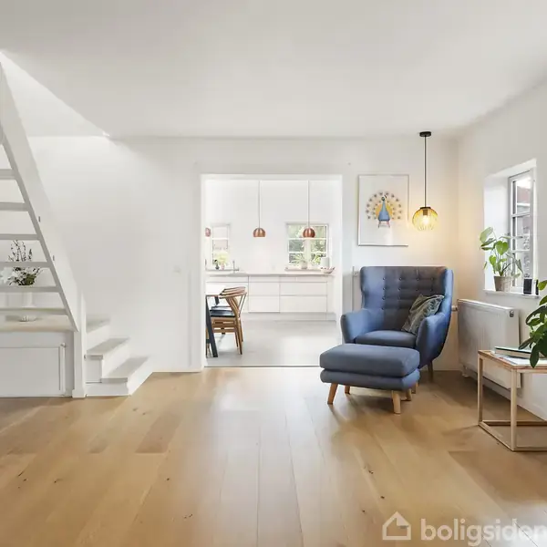 En moderne stue med trægulv, hvor en blå lænestol står ved et lille bord med planter. Trappen til venstre leder til en loftsetage. Køkkenet er synligt i baggrunden.