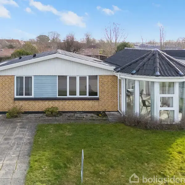 Et enetages gult murstenshus med et spidst tag er beliggende i et villakvarter. Vedhæftet er en glasveranda. Foran huset er en flisebelagt indkørsel og en velholdt græsplæne.