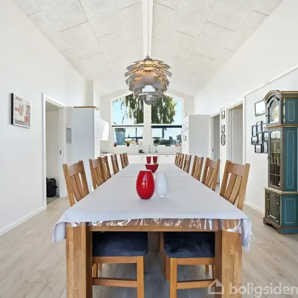 Langt spisebord med hvide dug og to røde vaser i midten. Omgivet af stole under en moderne lampe. Rummet har lyse vægge, høj loftshøjde og udsmykningen inkluderer kunst og et grønt vitrineskab.