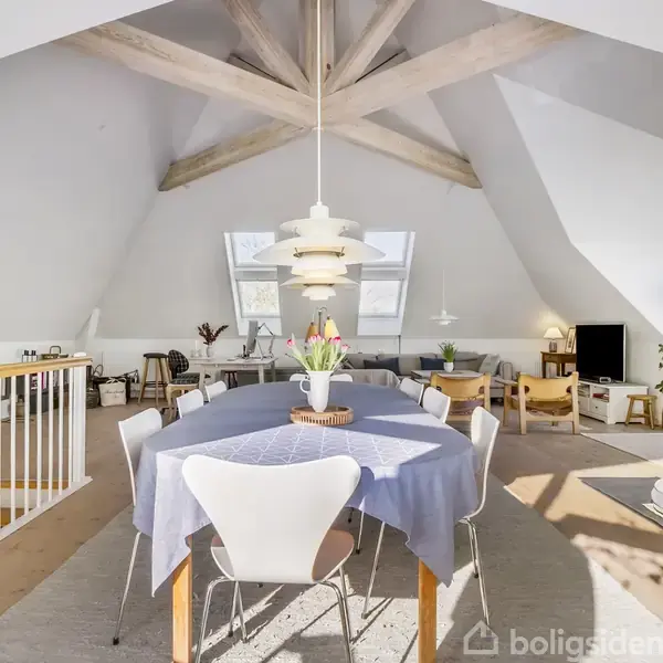 Et rummeligt spisestueområde med et rundt bord dækket med en blå dug og omgivet af stole. Rummet har en høj loftshøjde med synlige bjælker, store vinduer og lader naturligt lys strømme ind.