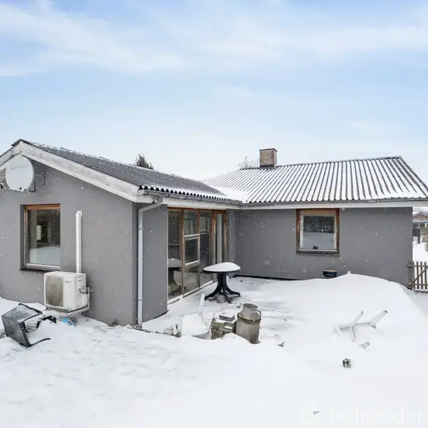 En brændeovn står tændt i en stue med trægulve og -loft. Til venstre ses et hvidt køkken med komfur og vinduer. En dør med glaspartier er til højre.