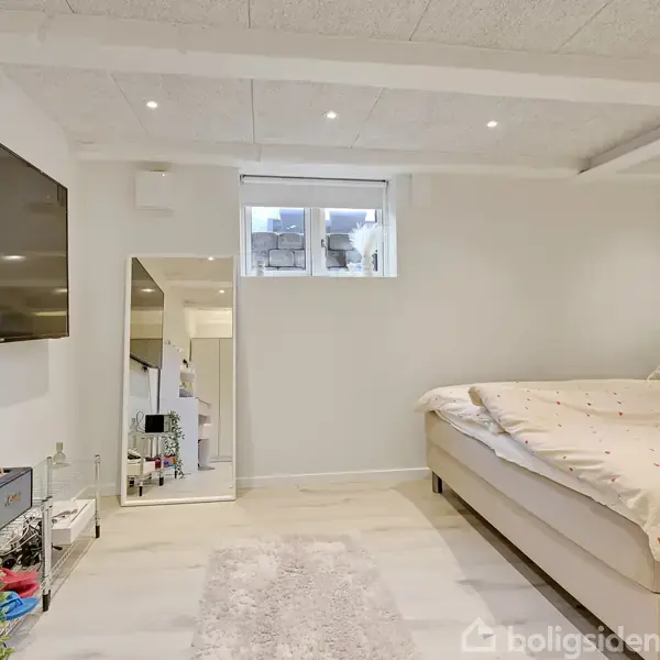 Et soveværelse har en stor seng til højre, et spejl ved siden af et lille vindue, og et tv hængende på væggen over en hylde med planter og opbevaring til venstre.