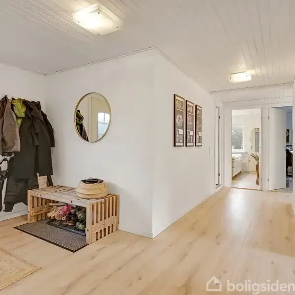 En lys gang med trægulv, hvor en bænk med kurv og overtøjshængere står til venstre. En rund spejl og flere billeder hænger på væggen. De leder mod åbne værelser.