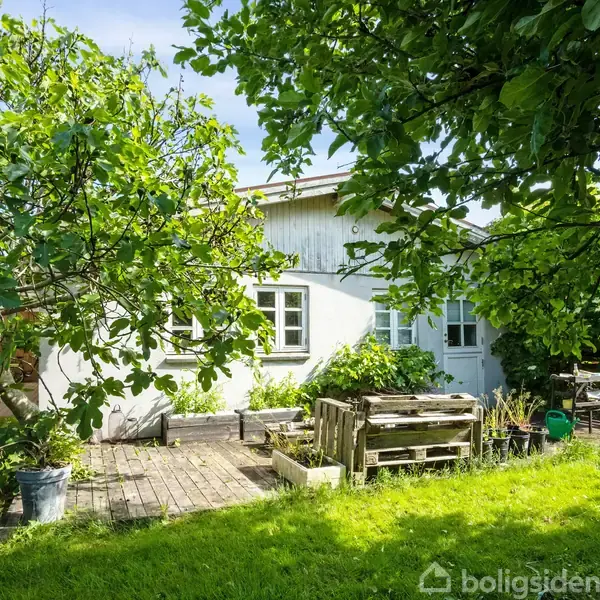 Et hvidt hus med hvide vinduer står i en frodig have. Foran huset er der en terrasse med træmøbler omgivet af grønne træer og græsplæne.