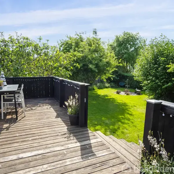 Træterrasse med sorte møbler står på en solrig dag. Terrassen er omgivet af frodig have med grønne træer og buske. I baggrunden ses en velplejet græsplæne med havemøbler.
