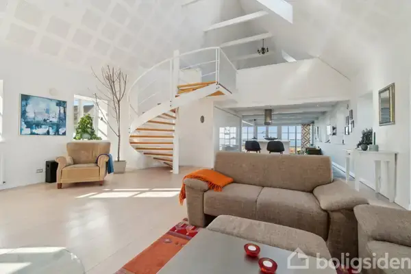 Stue med beige sofaer står i forgrunden, orange tæppe og trappe til første sal bagved. Lyse trægulve og store vinduer giver et moderne, lyst udtryk.