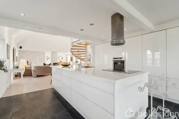 Et moderne køkken med en stor, hvid ø og indbygget komfur, omgivet af en åben stue med en vindeltrappe i baggrunden. Indretningen er stilren med lyse farver.