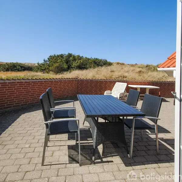Et sort havebord med flere stole står på en flisebelagt terrasse omgivet af en lav mur. Terrassen har udsigt til klitter og blå himmel på en solrig dag.