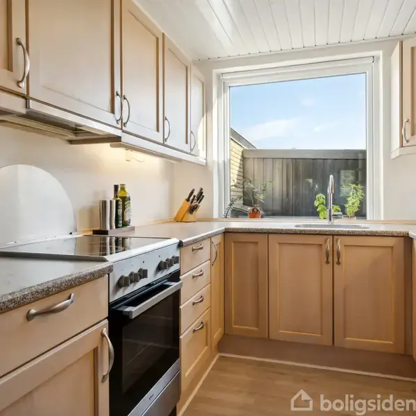 Et køkken med træelementer og en sort komfur står i hjørnet. Overfladerne har køkkenredskaber, og et stort vindue giver udsigt til en træhegn og plante udenfor.