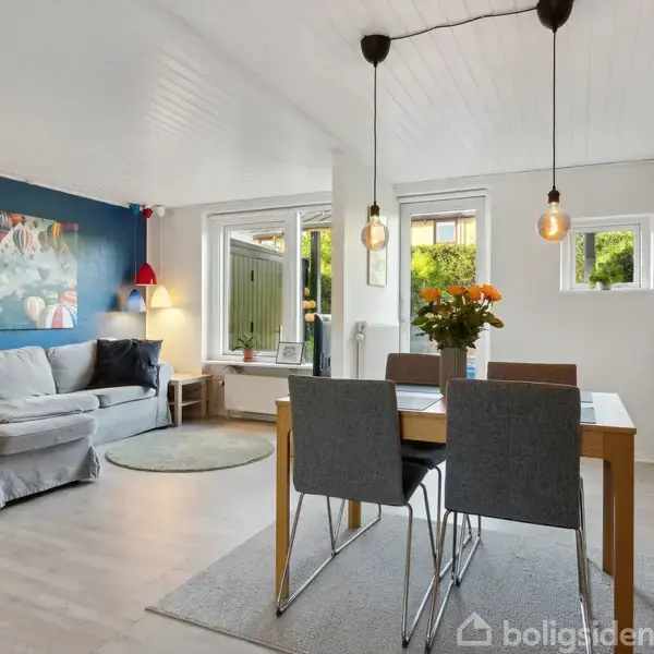 En stue med en grå sofa og farverigt billede på en blå væg. Et spisebord er dækket med blomster og omgivet af stole, mens store vinduer giver naturligt lys.