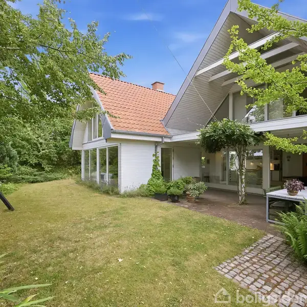Et moderne hus med store vinduer og et fladt tag udført i træ og tegl beliggende i en grøn have med en gyngestativ i baggrunden.