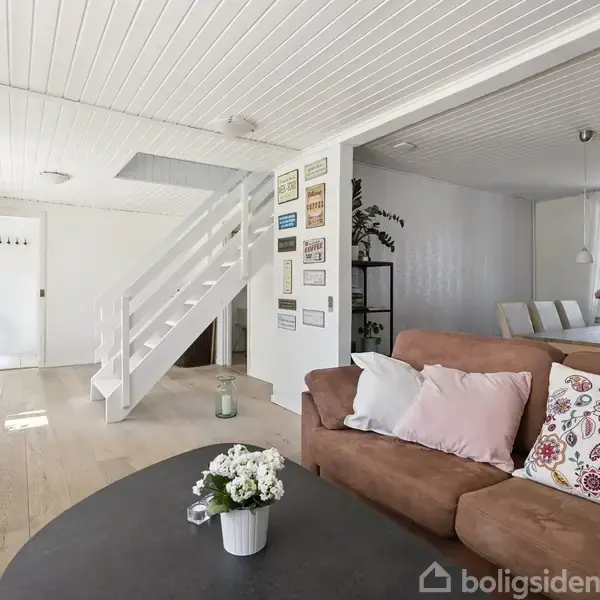 Brun sofa med puder står i stue. Stuen har trægulv, hvide vægge og et hvidt loft. Trappe fører opad. Blomsterkrukke på bordet. Beskedplader hænger på væggen.