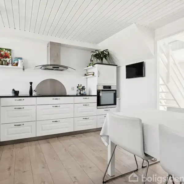 Lyst køkken med hvide skabe og sort bordplade, en ovn indbygget i højre skabsvæg. Bord med to stole ved vindue, og planter på bord og hylder. Trægulv og hvidt loft.