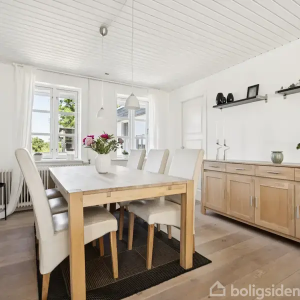 Spisebord med hvide stole omkring, står i et lyst rum med trægulv. Vinduer med lysgardiner bagved, en skænk med planter og dekorationer til højre.