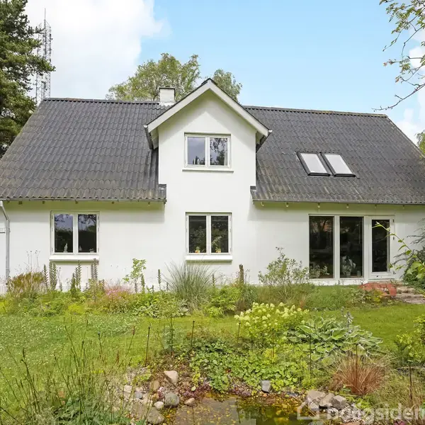 Hvidt hus med sort tag står i en frodig have med græs og forskellige planter. To store vinduer og en havedør vender ud mod haven.