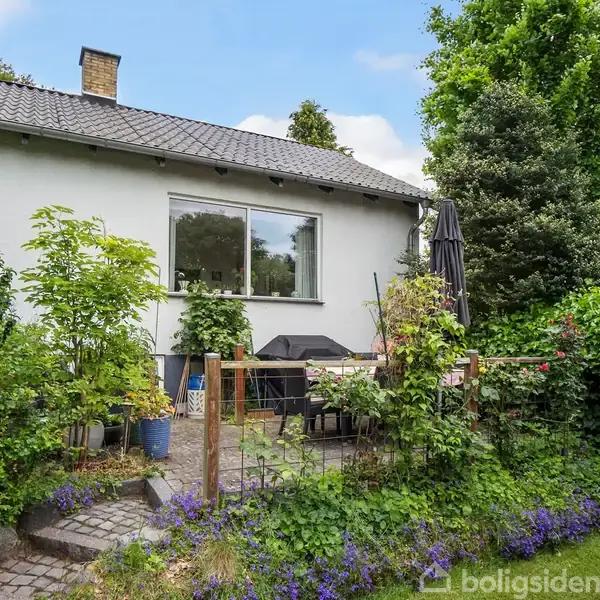 Hus med hvid facade og skråt tag. En terrasse foran huset har havemøbler og en parasol. Omgivet af frodig have med grønne planter og blomster.