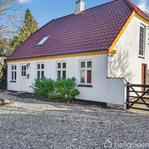 Hvidt hus med rødt tag står stille på grusbelagt grund. Omkring huset er der træer og en træport. Sollys skaber skygger på husets facade.
