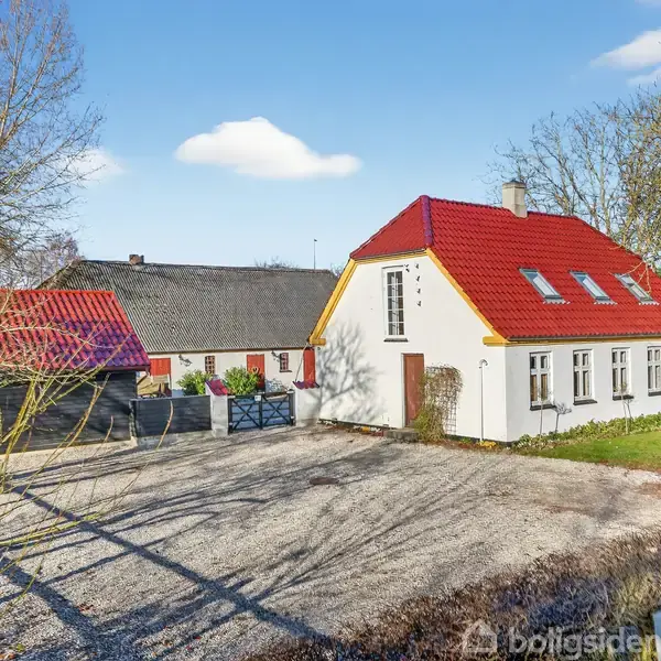 Hus med rød tag og hvide vægge står på en gårdsplads med grus, omgivet af træer og en gammel lade i baggrunden.