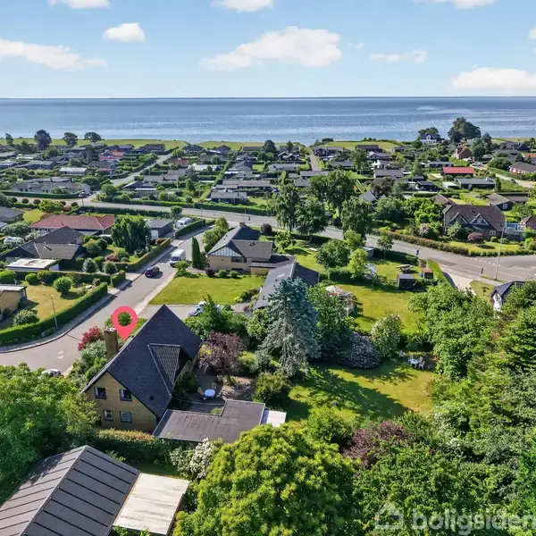 En stor villa med skråt tag ligger i en frodig have. Omkringliggende huse og veje ses i forstaden. I baggrunden breder en stor vandmasse sig under en blå himmel.