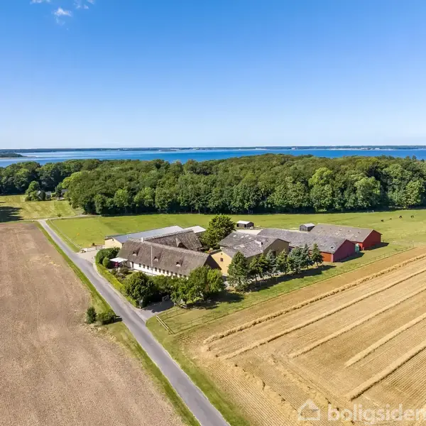 En landejendom omgivet af marker i høst. Bygningerne står tæt ved en skov, med havet i baggrunden under en klar blå himmel.