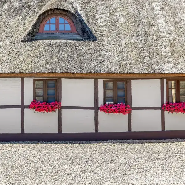 Stråtækt bindingsværkshus med fire vinduer dekoreret med røde blomsterkasser. Grusbelagt gårdsplads foran huset, som udstråler en rustik og traditionel atmosfære.