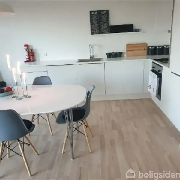 Et hvidt spisebord med sorte stole står i et lyst køkken. På bordet er der en lysestage med tændte stearinlys og en skål med frugt. Køkkenet har hvide skabe.