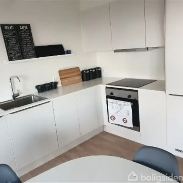 Et moderne køkken med hvide skabe rummer en vask, skærebræt og kogeplader. Der er tavler over bordpladen med teksten "GELATI" og "CAFFÈ". Indretningen er minimalistisk og lys.