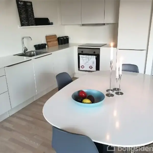 Et moderne køkken med hvidt spisebord og fire stole. En blå skål med frugter og to tændte stearinlys står på bordet. Ovn, håndvask og skab omkranser rummet.