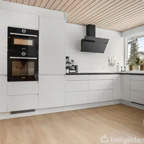 Hvidt, moderne køkken med indbyggede ovne og sorte kogeplader. En emhætte er monteret over kogepladen. Køkkenudstyr og planter står på bordpladen. Rummet har trægulv og træloft.