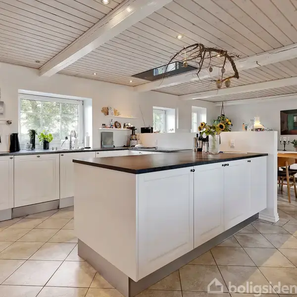En moderne køkkenø med hvide skabe står i midten af et lyst køkken. På øen er der blomster. Omkring er der store vinduer, træloft og spiseområde med spisebord og stole.