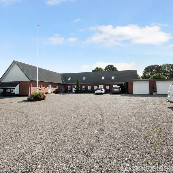 En stor, murstensbygning med mørkt tag står på en grusbelagt plads. Flere biler er parkeret foran bygningen, omkranset af træer og blå himmel.