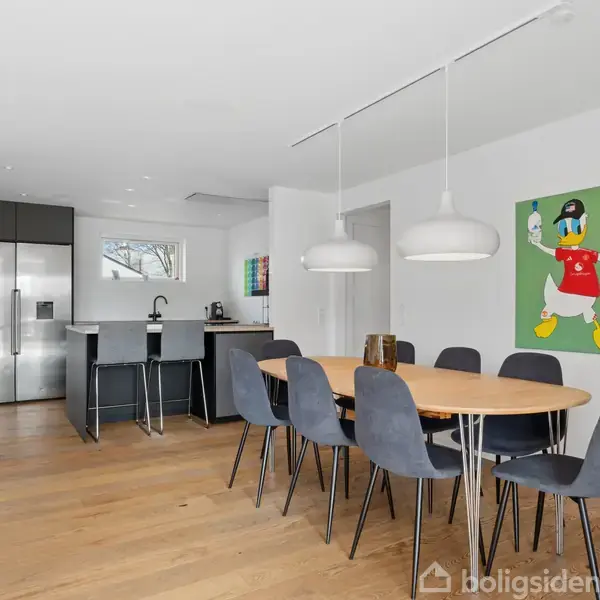 Et spisebord med otte stole står i et moderne køkken. Køkkenet har en stor metal-køleskab og en ø med barstole. På væggen hænger et maleri af Anders And.