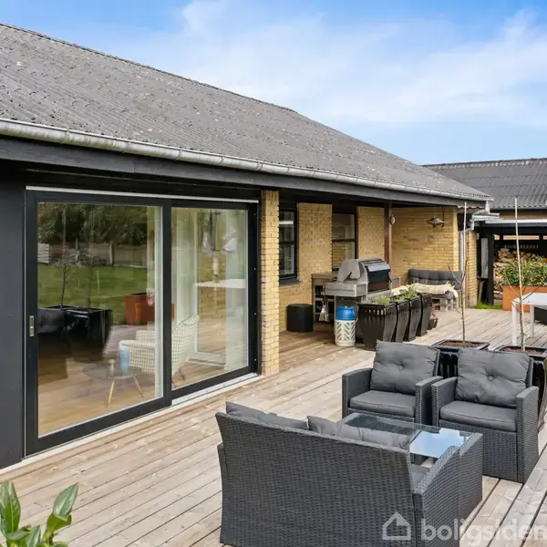 Rattanmøbler står på en rummelig træterrasse med grill ved siden af et murstenshus. Terrassen har en parasol, og gennem store vinduer ses ind i huset. Græsplæne omgiver området.