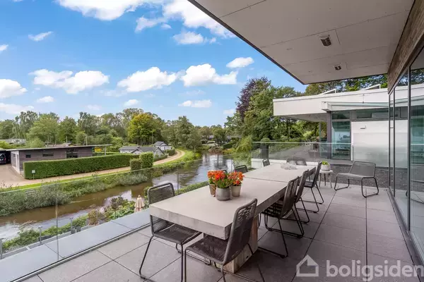 Lang altan med bord og stole står stille ved glasrækværk. Den omkranser et moderne hus ved en snoet flod omkranset af grønne træer. Solrig, klar himmel danner baggrunden.