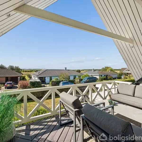 Terrasse med sofa og stole under et skråt tag, omgivet af et beboelseskvarter med huse og grønne områder, og med udsigt mod horisonten under en klar blå himmel.