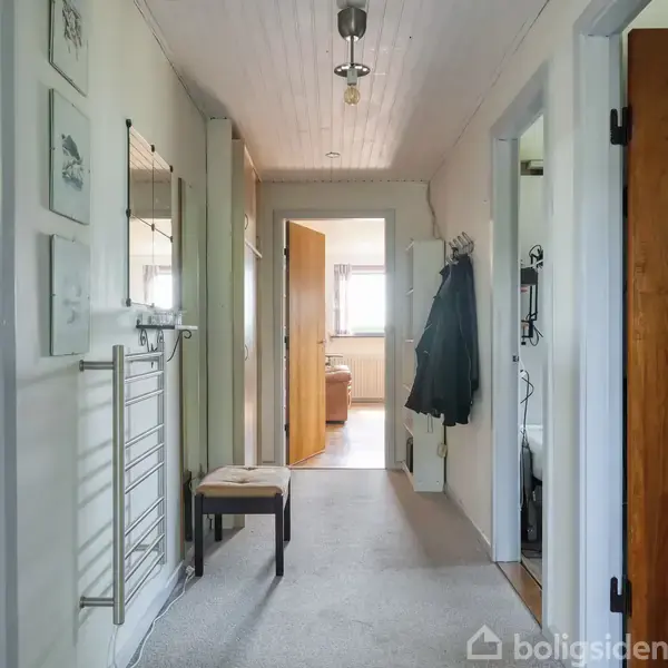 Gang med lyse vægge og loft. På væggen hænger billeder og en knagerække med jakker. Et spejl og en bænk er i venstre side. Døre fører til andre rum.