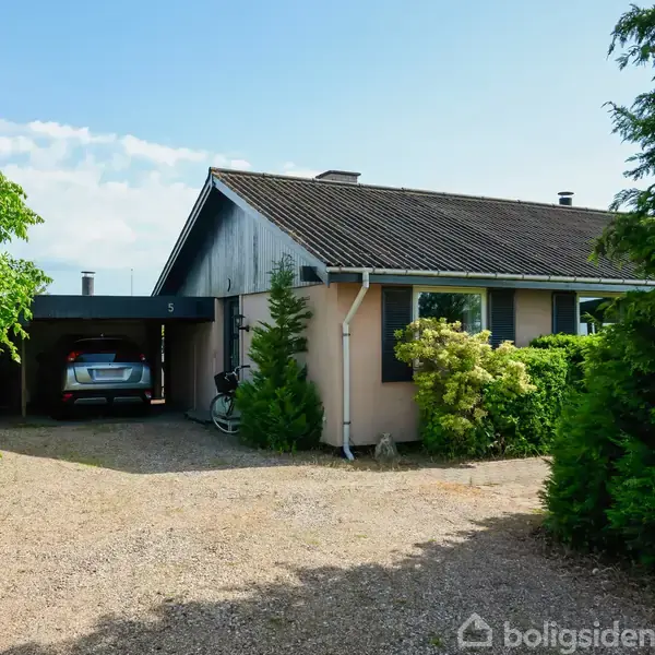 Et étplanshus med fladt tag og to store vinduer. En bil holder i en carport til venstre. Omgivet af tæt, grøn beplantning og grus indkørsel. Husnummer 5 ses.
