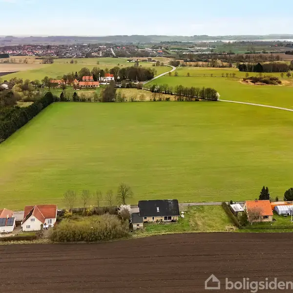 Flere huse ligger langs en vej ved kanten af en stor grøn mark, omgivet af landligt landskab med marker og skove i baggrunden.