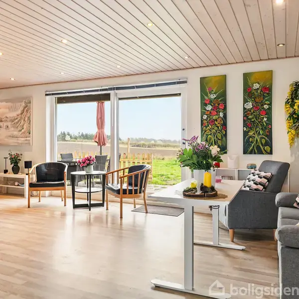 Stue med møbler: en sofa, stole og et bord. Vaser med blomster står på bordene, og store vinduer åbner op til en græsplæne. Malerier pryder væggene.