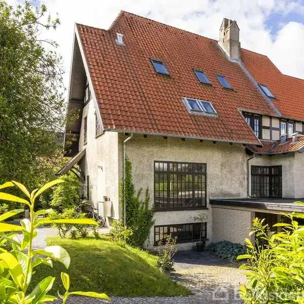 Et stort hus med rødt tegltag og flere vinduer. Det er omgivet af en grøn have med buske og træer. En brostensbelagt sti fører op til huset.