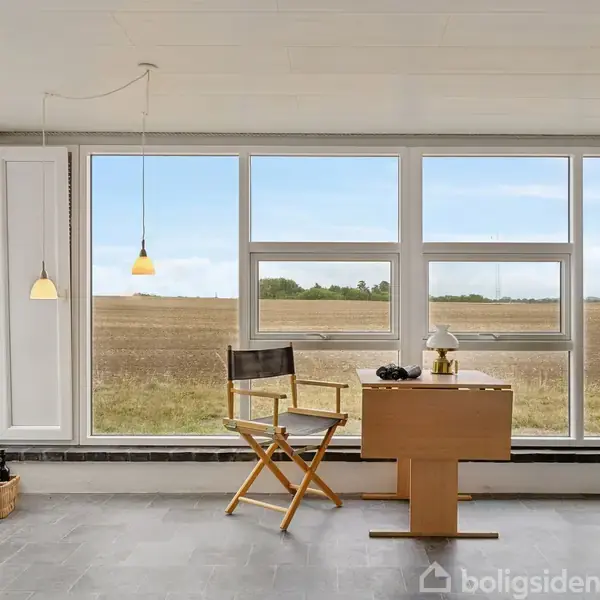 Et bord og en stol står i et lyst rum med store vinduer, der giver udsigt til en mark. To lamper hænger fra loftet, og en kurv står til venstre.