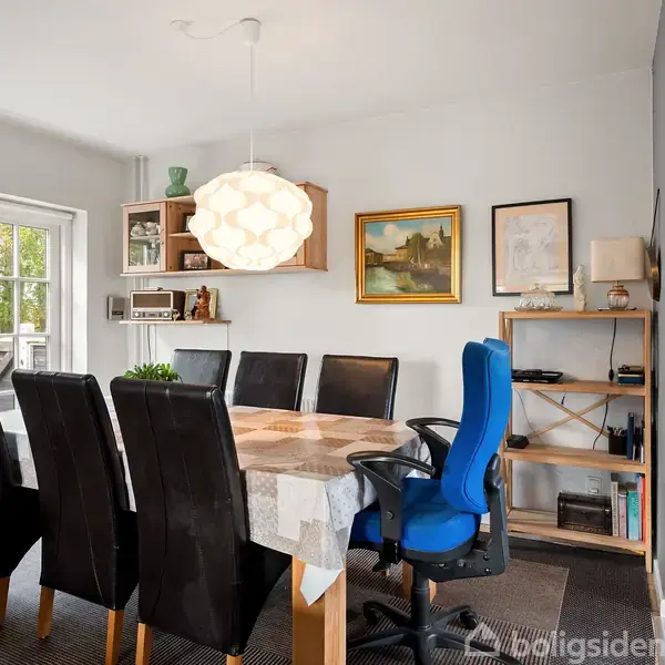 En spisebord med seks sorte stole og en blå kontorstol står under en hængelampe. Bagved ses en væghylde med dekorationer og billeder, nær en stor glasdør til venstre.